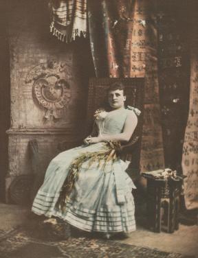 A portrait of a woman in a gown, sitting in a chair against a backdrop of curtains and blankets.