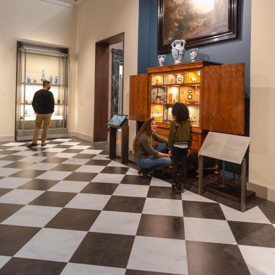View of gallery with parent kneeling by large wooden dollhouse cabinet with multiple rooms visible; parent looking up at child standing adjacent