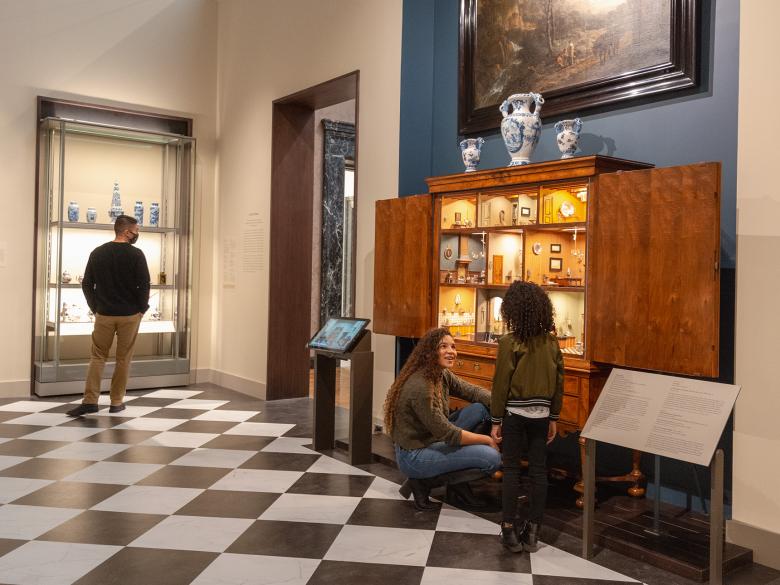 To the right, a visitor looks at a glass case full of blue-and-white ceramics. To the left, a visitor with a young child looks at an ornate doll's house.