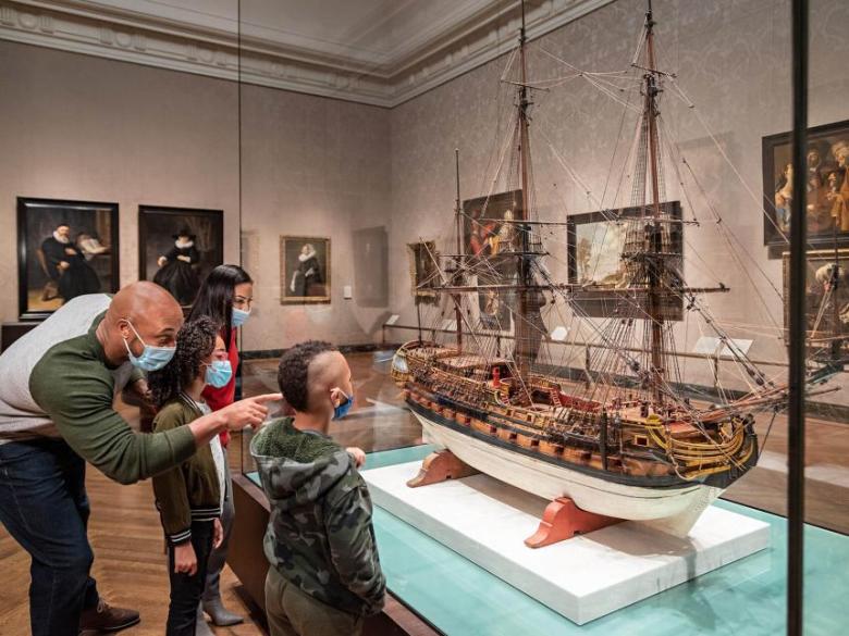 A visitor with two young children looks at a Dutch model ship behind a glass case.