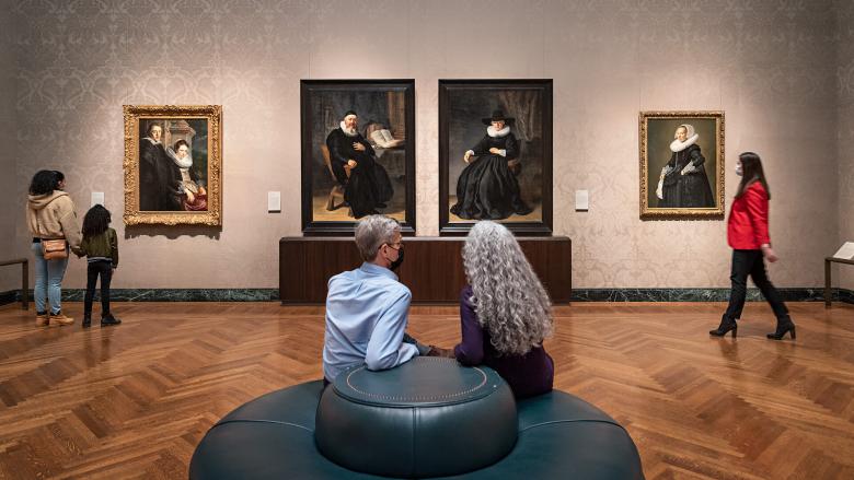 Two visitors sitting on couch in middle of gallery with Dutch portraits hanging on walls; other visitors walking by paintings
