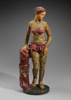 A small papier mache statuette of a heavily tattoed woman in a red bathing suit stands on a pedestal and holds a matching red garment with a crest that reads "Battleship Kate."