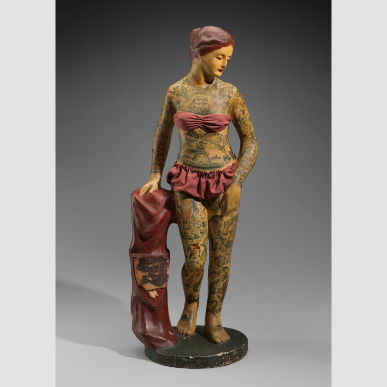 A small papier mache statuette of a heavily tattoed woman in a red bathing suit stands on a pedestal and holds a matching red garment with a crest that reads "Battleship Kate."