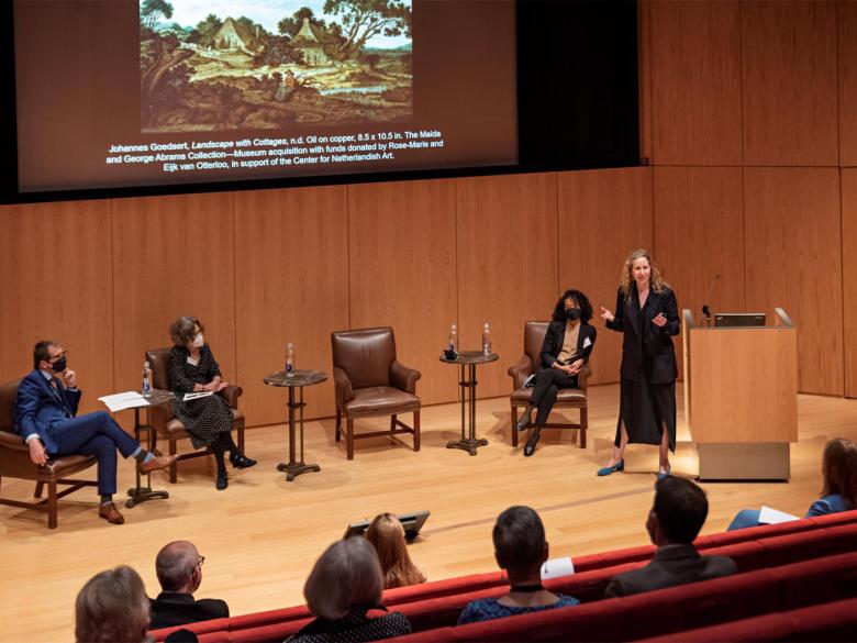 A person stands on a stage in front of a screen showing a Flemish art work and addresses an audience.