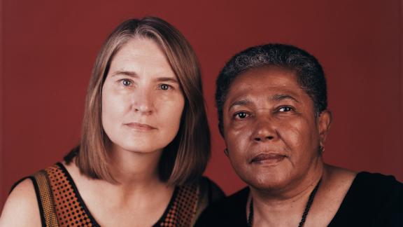 Two individuals shown from the chest up sit closely next to each other and pose for a photo against a red background.