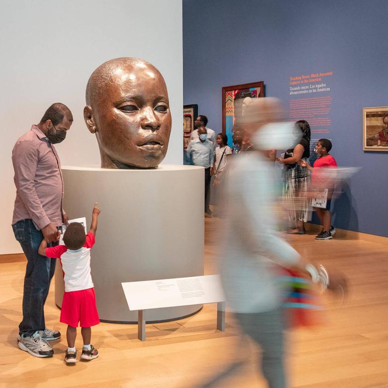 Various visitors in gallery; parent and child, pointing up, looking at large sculpture of bald baby head
