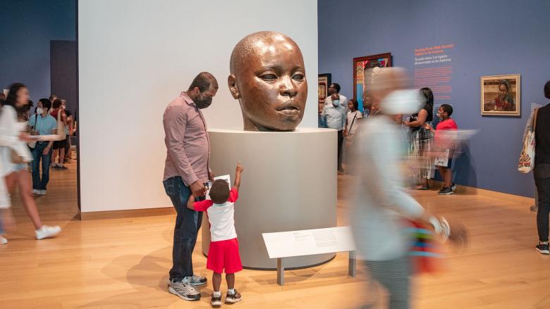 Various visitors in gallery; parent and child, pointing up, looking at large sculpture of bald baby head