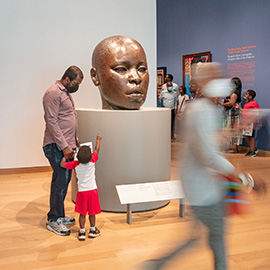 Various visitors in gallery; parent and child, pointing up, looking at large sculpture of bald baby head