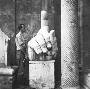 Cy Twombly standing aside a large stone hand with the index finger pointing upwards. 