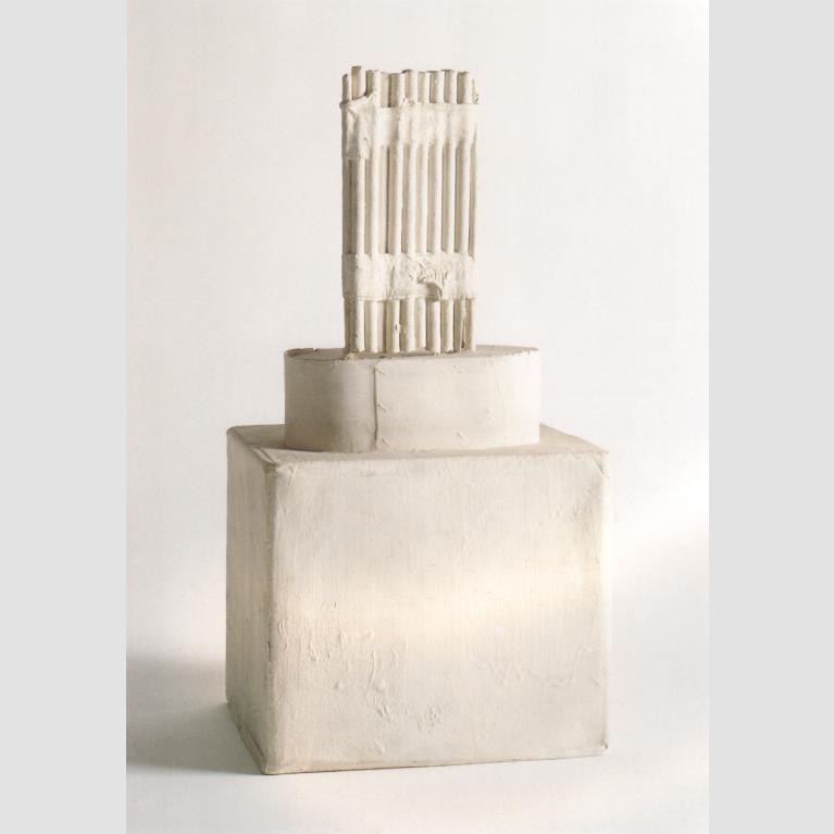A sculpture of bundled sticks placed on top of a cylinder sat atop a box. 