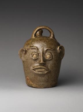 An earthenware jug with a small opening, a handle, and decorated with the features of a human face.
