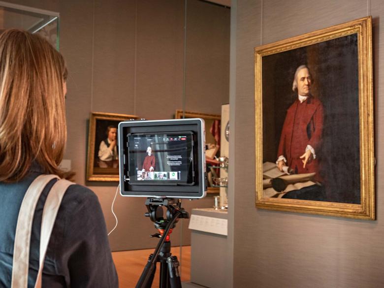 An MFA instructor operating a camera on a tripod pointed at portrait painting of Samuel Adams by Copley