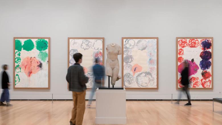 Visitor standing in foreground looking at ancient Greek sculpture on pedestal, with other visitors in background looking at Twombly paintings hanging on wall