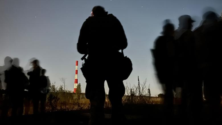 The silhouettes of Ukrainian soldiers stand out against the evening sky; a fluorescent orange and white tower sits in the distance. 