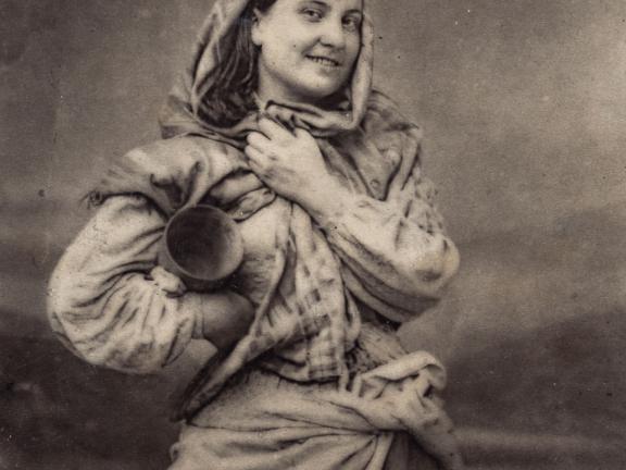 A young woman smiles for the camera with a cloth wrapped around her head and her right arm akimbo, carrying a jug. 