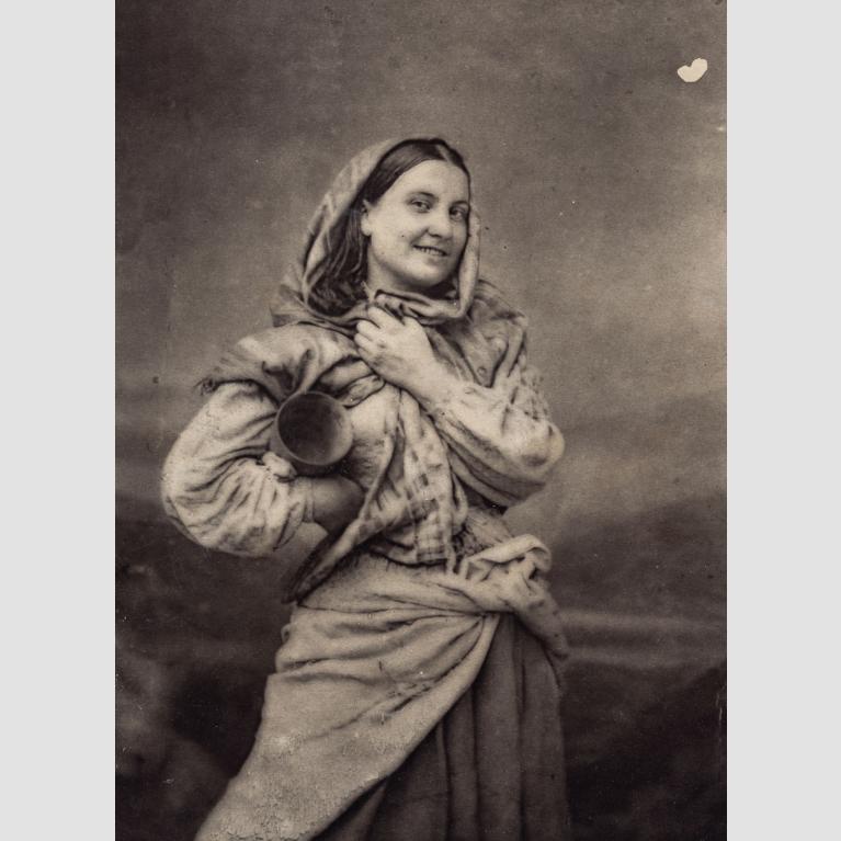 A young woman smiles for the camera with a cloth wrapped around her head and her right arm akimbo, carrying a jug. 