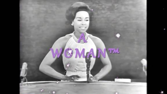 A woman in a gown with arms akimbo stands in front of three microphones.