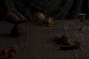 A lace-covered table set with pigs feet on a cake stand, moldy fruit and vegetables, and a goblet. 