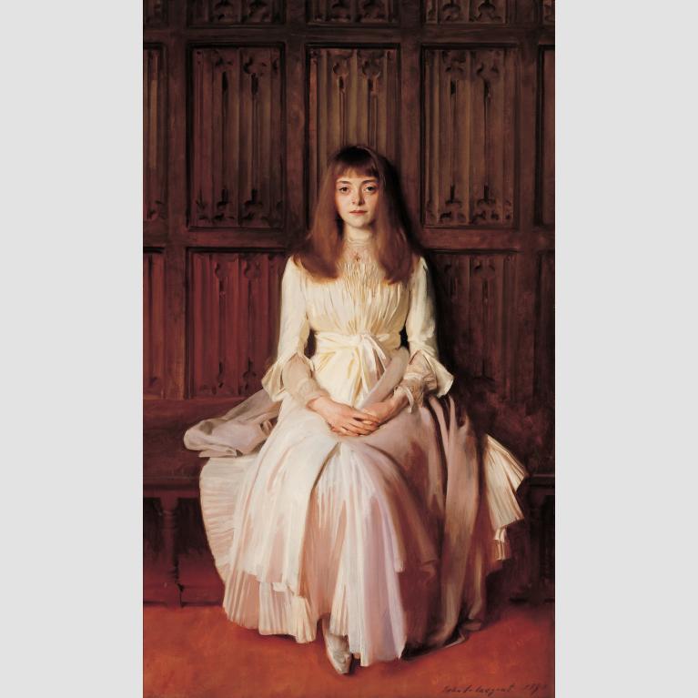 A young girl in a pale gown sits on a wooden bench with her hands clasped on her lap.