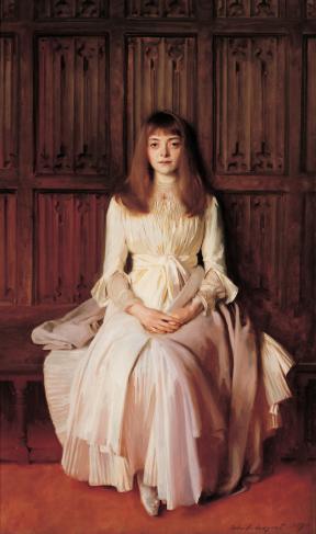 A young girl in a pale gown sits on a wooden bench with her hands clasped on her lap.