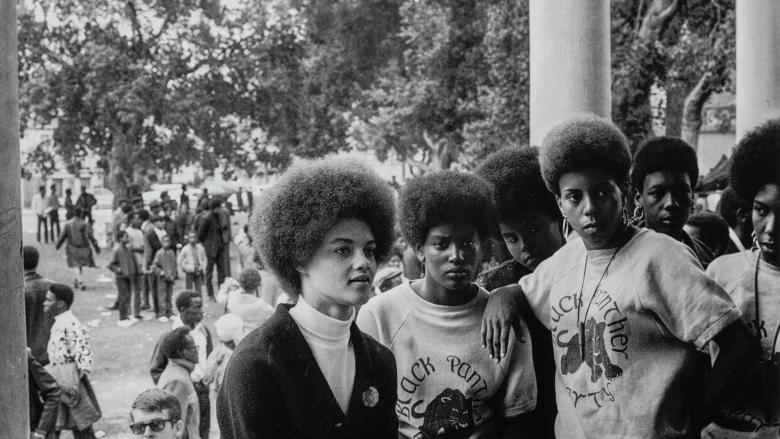 Black-and-white photo: a group of Black women look off into the distance as they stand before a crowd gathered on a lawn.