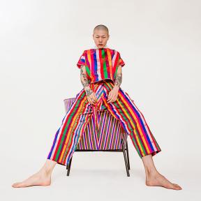 A model in a matching rainbow-striped shirt and pants leans against a piece of furniture covered in a fabric that matches his outfit.