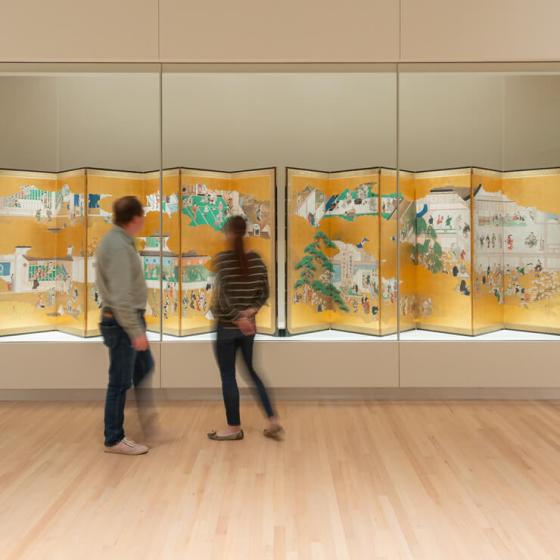 Two visitors looking at pair of Japanese screens depicting a town and its many citizens