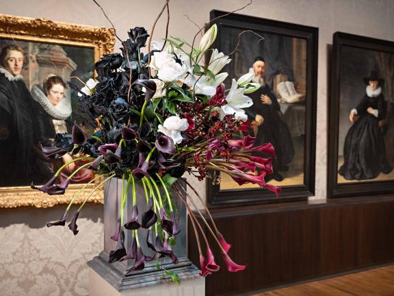 A large bouquet of black, red, and white flowers in front of several Dutch oil paintings 