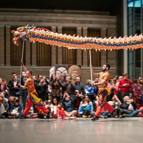 Two performers dressed in traditional Chinese outfits running by crowd in Shapiro Family courtyard, holding sticks supporting long dragon