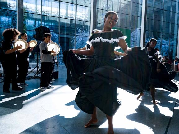 A woman in a black dress dances.  She is surrounded by other people playing hand percussion instruments.