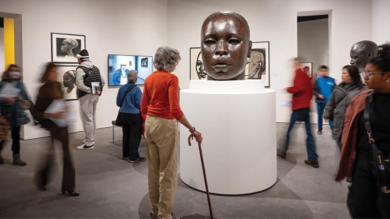 Visitor holding cane looking at large sculpture of bald head on tall white pedestal, with other visitors walking around in the gallery and looking at drawings on walls