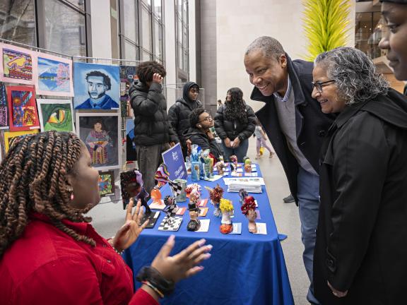 A small group of people gather around a young artist who sits next to a display of various artworks on a table and hanging behind her