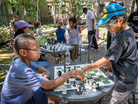 Two children sit in a sunlit courtyard and play chess.