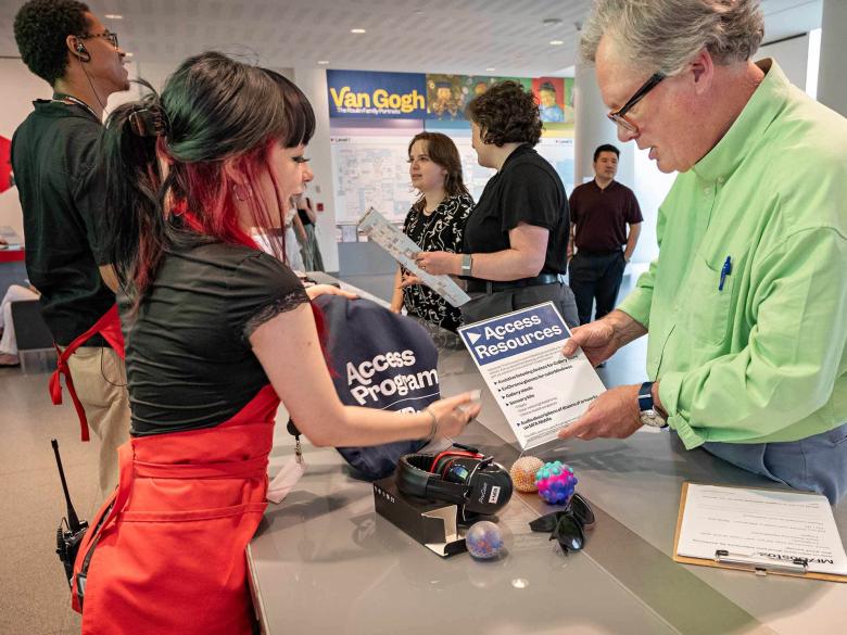 MFA Ambassador in red apron handing dark blue bag labeled Access Programs to visitor in green shirt at Sharf Visitor Center desk