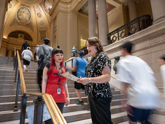 MFA Ambassador in red apron standing on staircase looking at Museum map with visitor