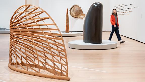 Visitor in red jacket walks around tall, finger-like, black stone sculpture, with wooden trellis-like sculpture in foreground