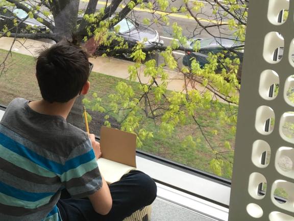 Teen sitting by window, holding sketchpad, looking out to grassy area