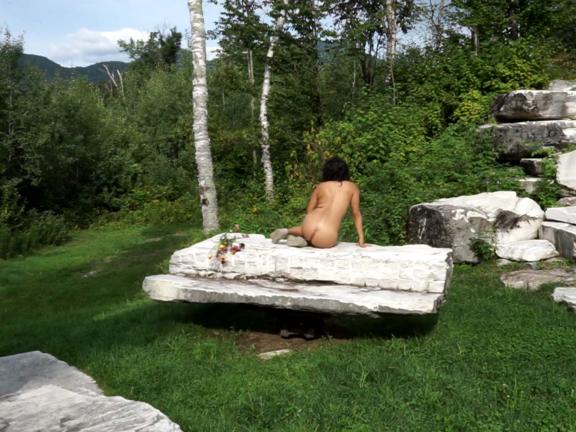 Video still depicting back of nude woman on stone platform in forest setting