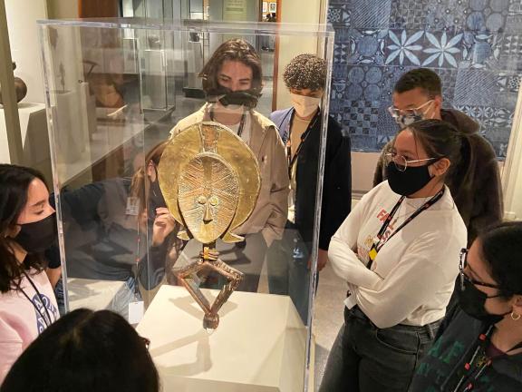 Teens wearing masks looking at and standing around glass case containing metal sculpture featuring a figure with a large headdress and bent knees